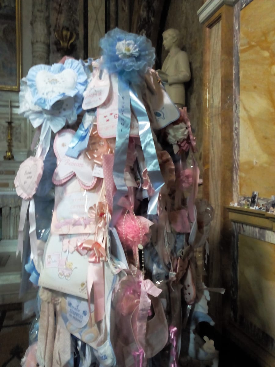 The marble chapel of St. Dominic Savio in the Basilica of Mary Help of Christians at Turin, with tiers of pink and blue ribbons, votive lamps, and a glass casket beneath the altar.