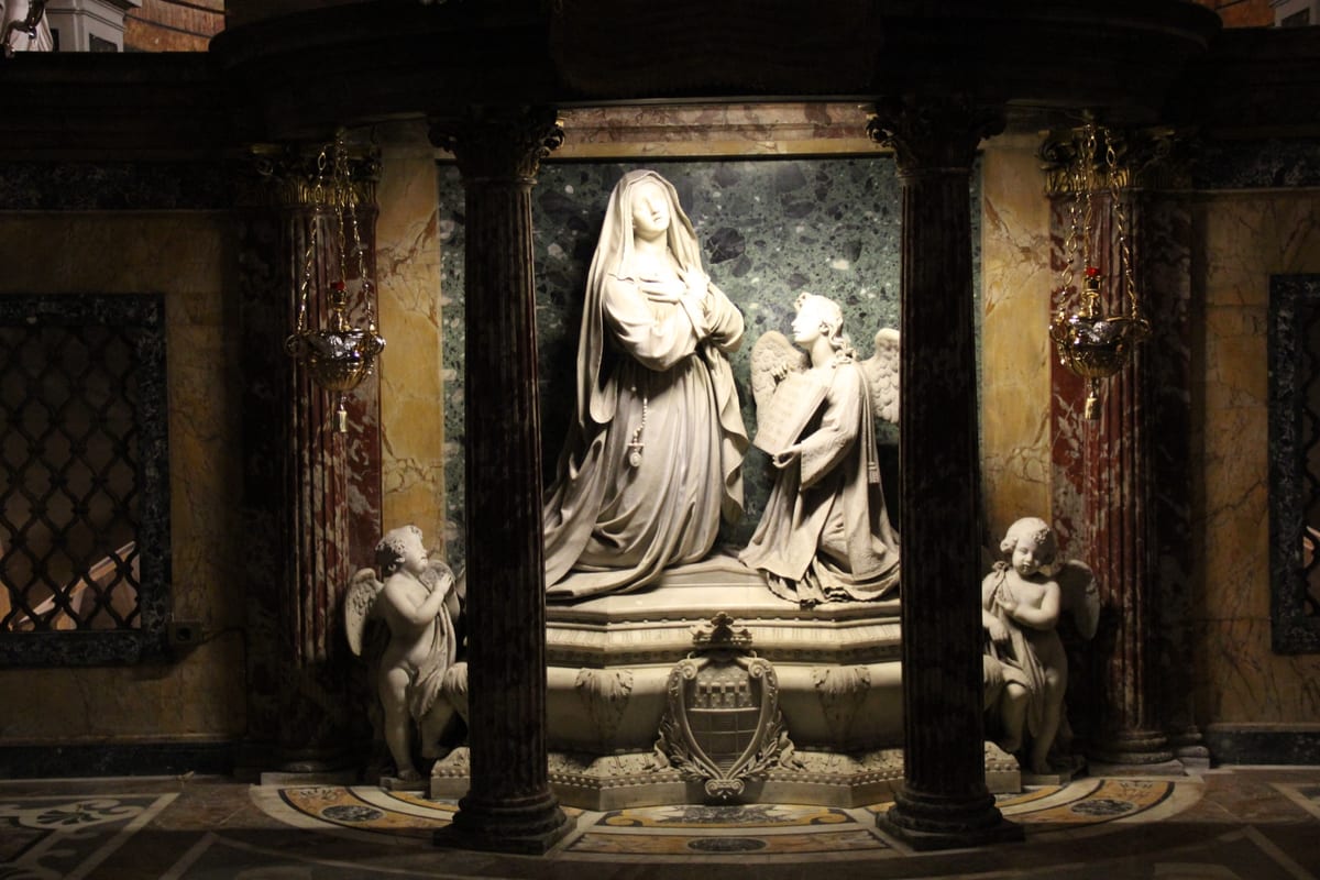 A white marble statue of St. Frances of Rome kneels with her hands crossed on her breast between dark red marble columns, beside a kneeling angel who holds an open book up to her.