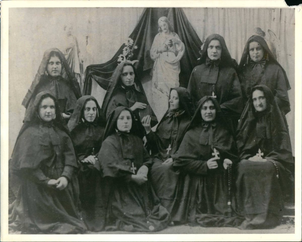 A formal portrait of Mother Cabrini seated with ten of her Missionary Sisters of the Sacred Heart of Jesus in habit and veil, arranged in two rows before a draped image of the Sacred Heart of Jesus.