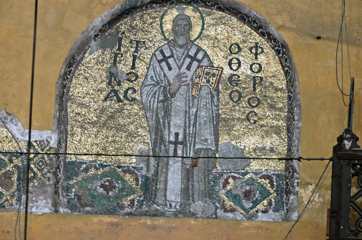 A Byzantine gold-ground mosaic shows St. Ignatius as an old, grey-bearded bishop in white vestments, holding a jewelled Gospel book.