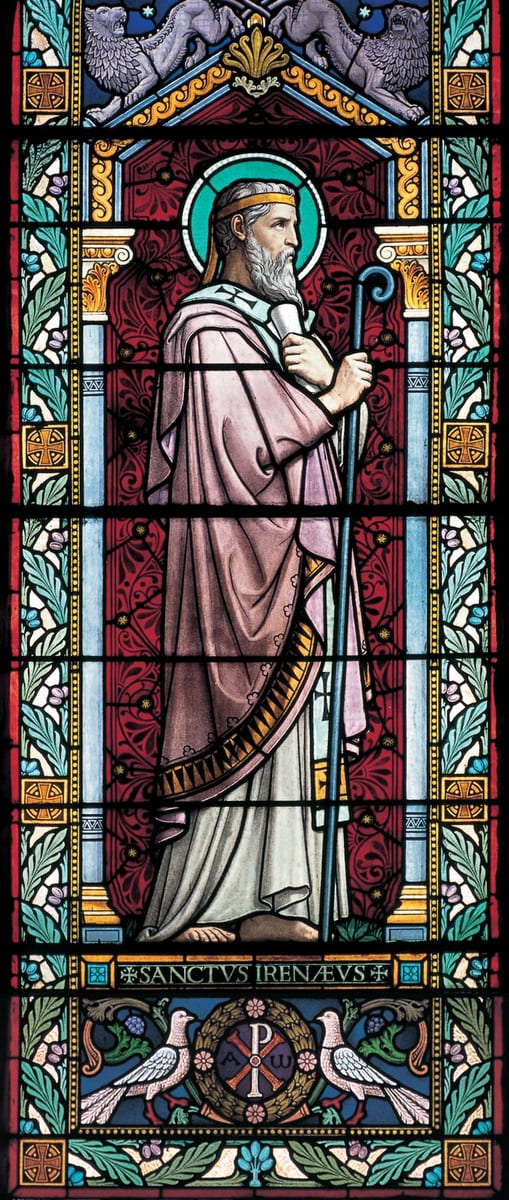 A stained-glass window depicting Saint Irenaeus as an elderly bearded bishop in profile, haloed, wearing a pink chasuble over a white and gold stole and holding a crozier, with the inscription SANCTVS IRENAEVS below.
