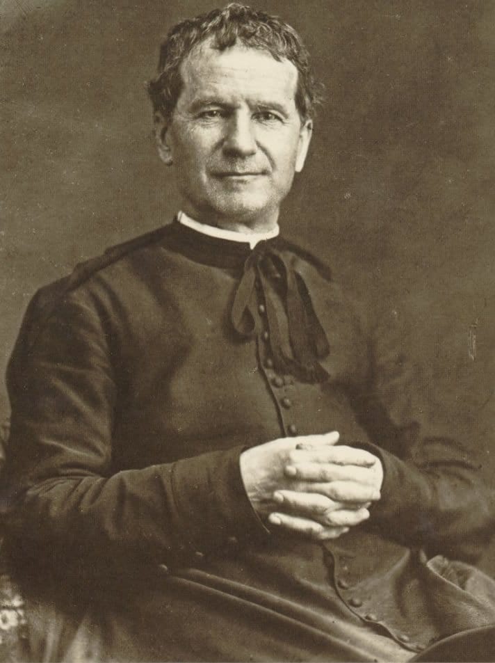 A seated man in a dark clerical cassock with a white collar and a dark ribbon at the throat sits with his hands clasped in his lap and looks steadily at the camera in an old sepia studio portrait.