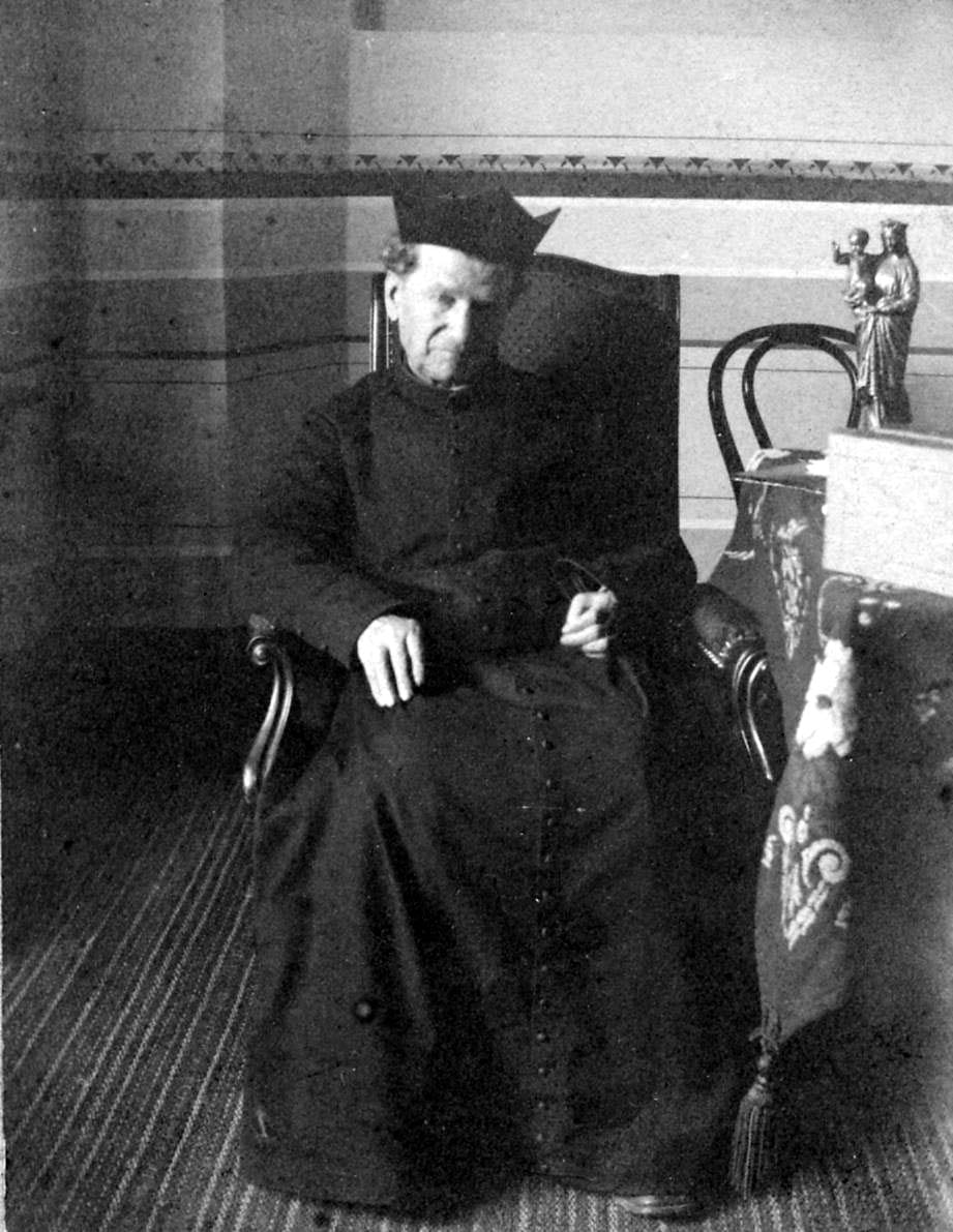Photograph of St. John Bosco in old age, seated in a bentwood armchair in biretta and cassock beside a table with a statuette of the crowned Virgin and Child.