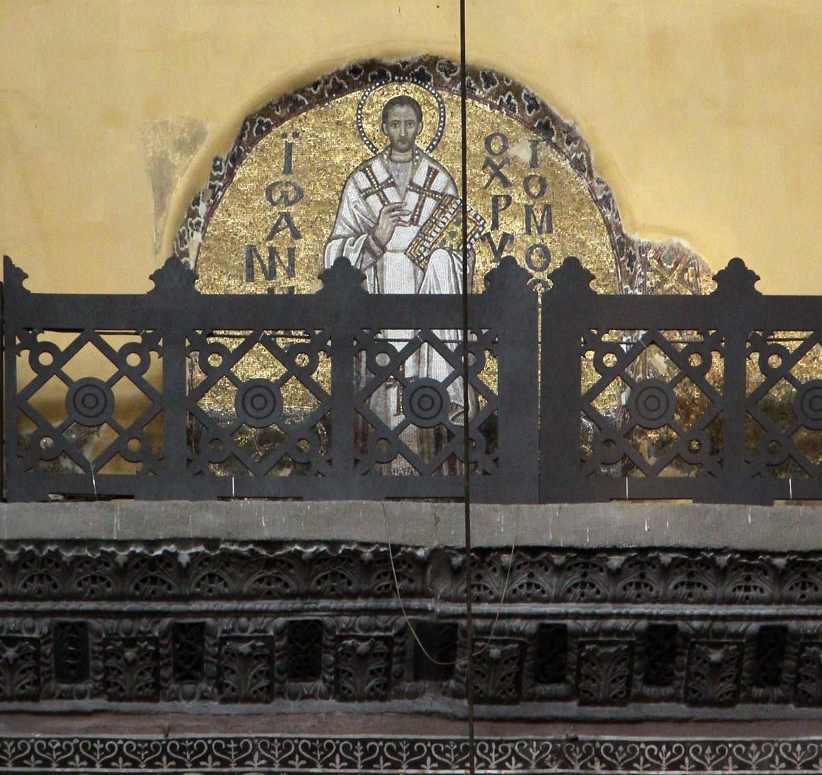 A gold-ground Byzantine mosaic in Hagia Sophia depicting Saint John Chrysostom as a bishop, haloed, vested in a white and cross-patterned omophorion, holding a jeweled Gospel book, with his name inscribed in Greek letters on either side.