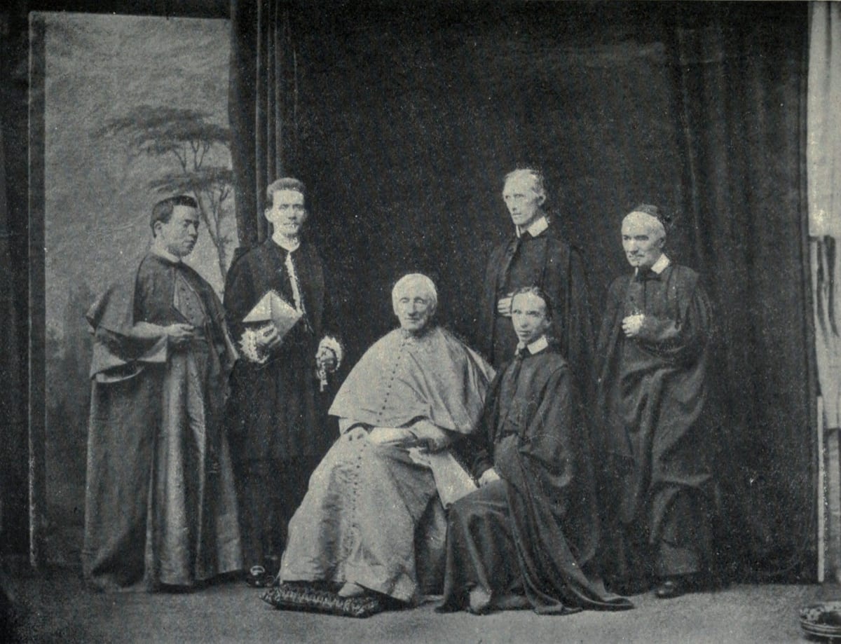 Cardinal St. John Henry Newman seated at the centre of a group of six in a Rome studio, in full choir dress, with three Oratorian priests and two attendants around him.