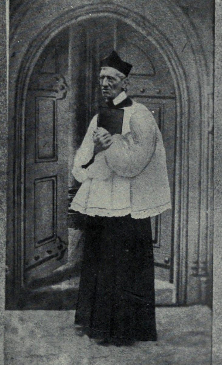 St. John Henry Newman standing in a Gothic church doorway before a heavy studded door, holding a small black book against his chest with both hands.