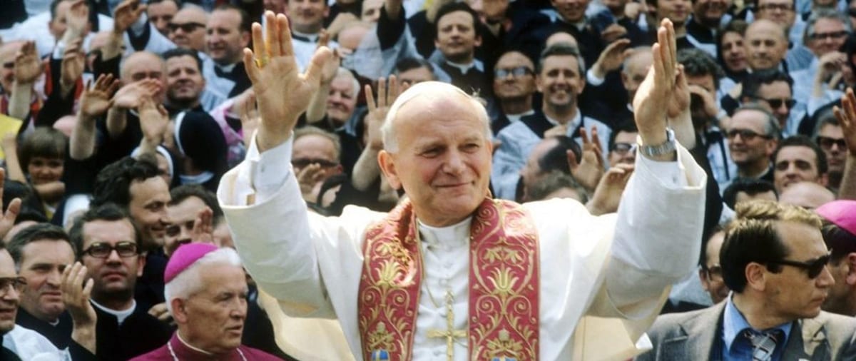 Pope John Paul II, in white cassock and red-and-gold stole, raises both hands to a cheering crowd of clergy and laypeople during his first pilgrimage to Poland in June 1979.