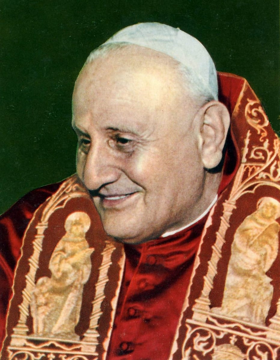 An elderly man in a white papal zucchetto and red papal vestments with gold-embroidered trim smiles warmly and turns his head to the left against a dark green background.