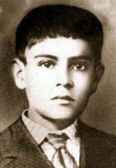 A studio portrait of a young dark-haired boy in a collared shirt and tie, looking directly at the camera.