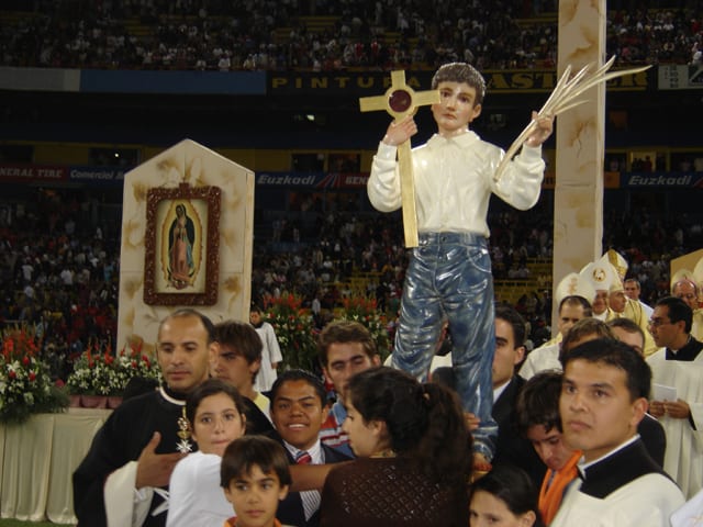 A statue of St. José holding up a wooden cross and a palm is carried above the crowd in the Estadio Jalisco at his beatification in 2005 AD.