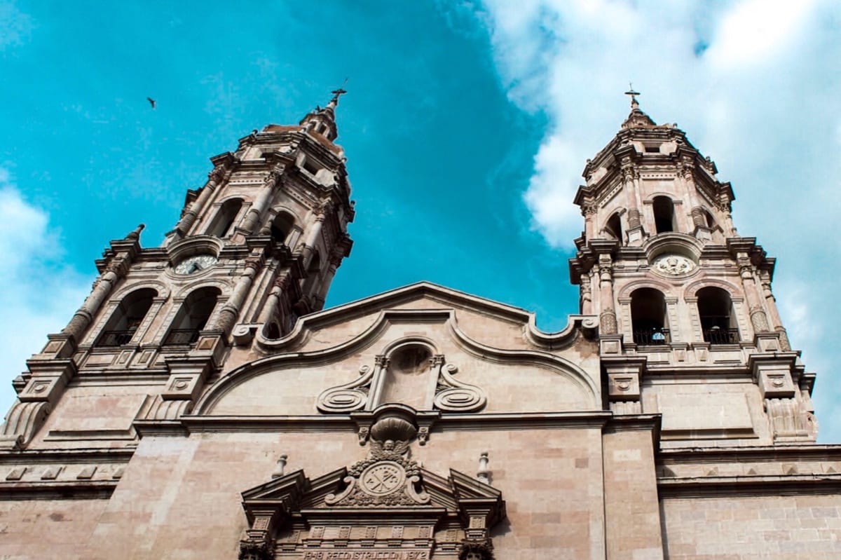 The twin-towered pink-stone front of the parish church of Santiago Apóstol in Sahuayo, as rebuilt between 1948 AD and 1972 AD, seen from below against a blue sky.