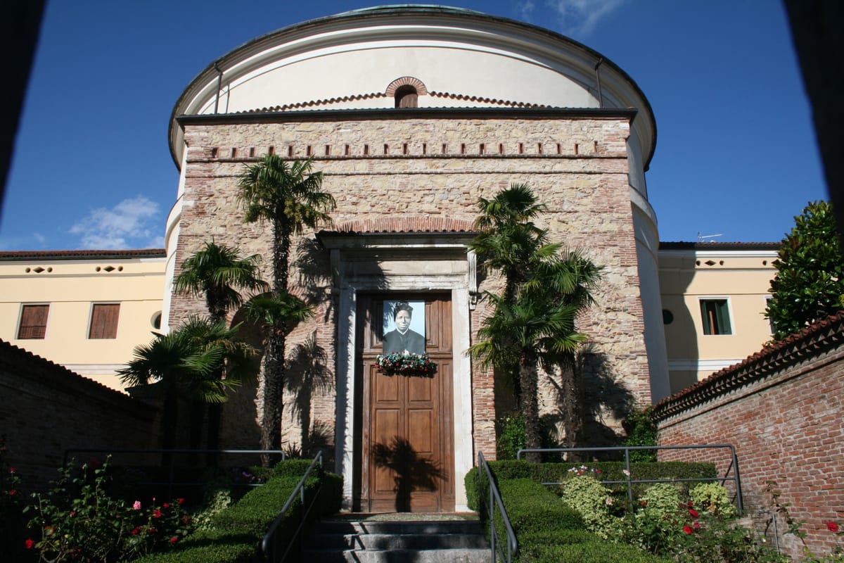 A colour photograph of the stone-and-brick front of the church of the Holy Family at Schio, with a large portrait of St. Josephine Bakhita hung over the door above a wreath of flowers.