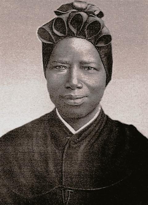 A black-and-white studio portrait of St. Josephine Bakhita, head and shoulders, smiling, in the black Canossian habit with a pleated bonnet and white collar band.