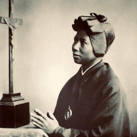 A sepia photograph of St. Josephine Bakhita in the Canossian habit, kneeling in profile at a draped prie-dieu before a tall wooden crucifix, hands joined.