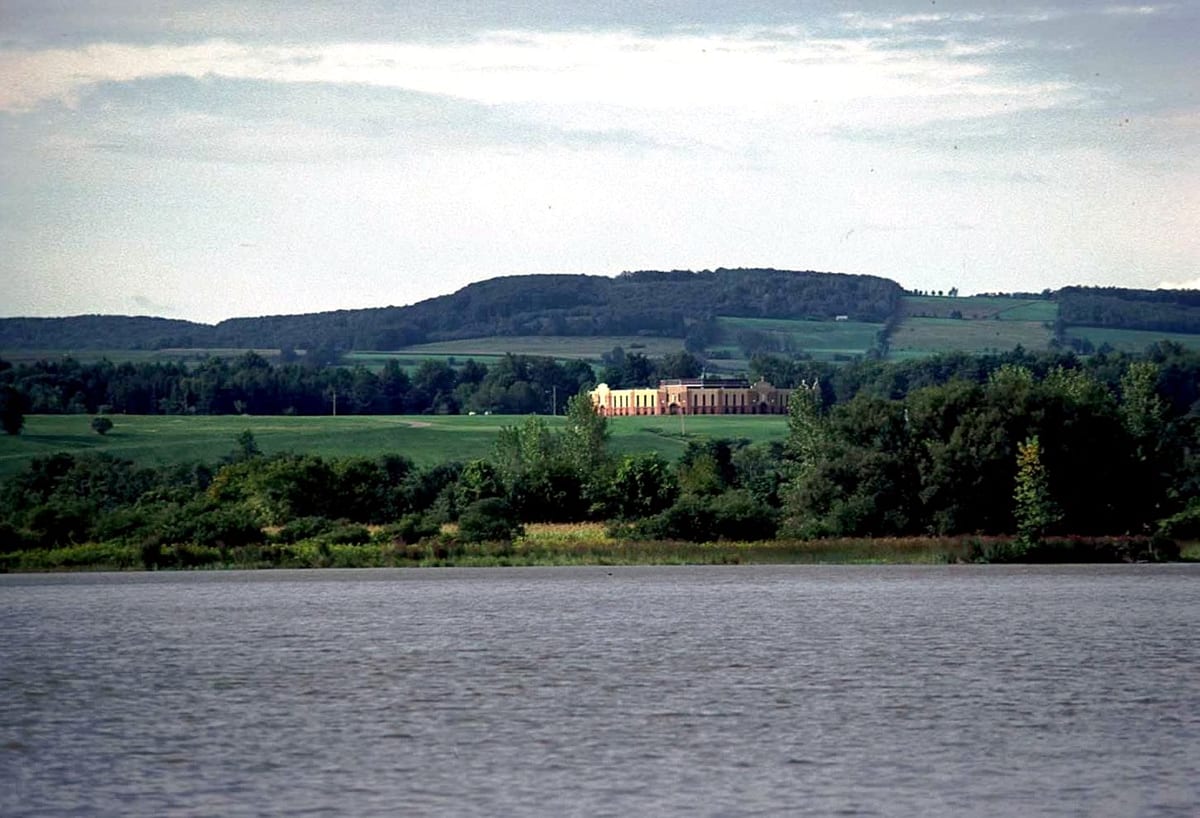 The Mohawk River at Auriesville, New York, with a reedy bank, trees, green hayfields and a long wooded ridge under an overcast sky.