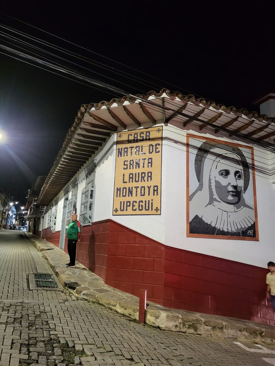 A white single-storey corner house with a clay-tile roof on a steep street in Jericó at night, with a yellow tile panel and a tile mosaic of St. Laura's face in the habit beside it.