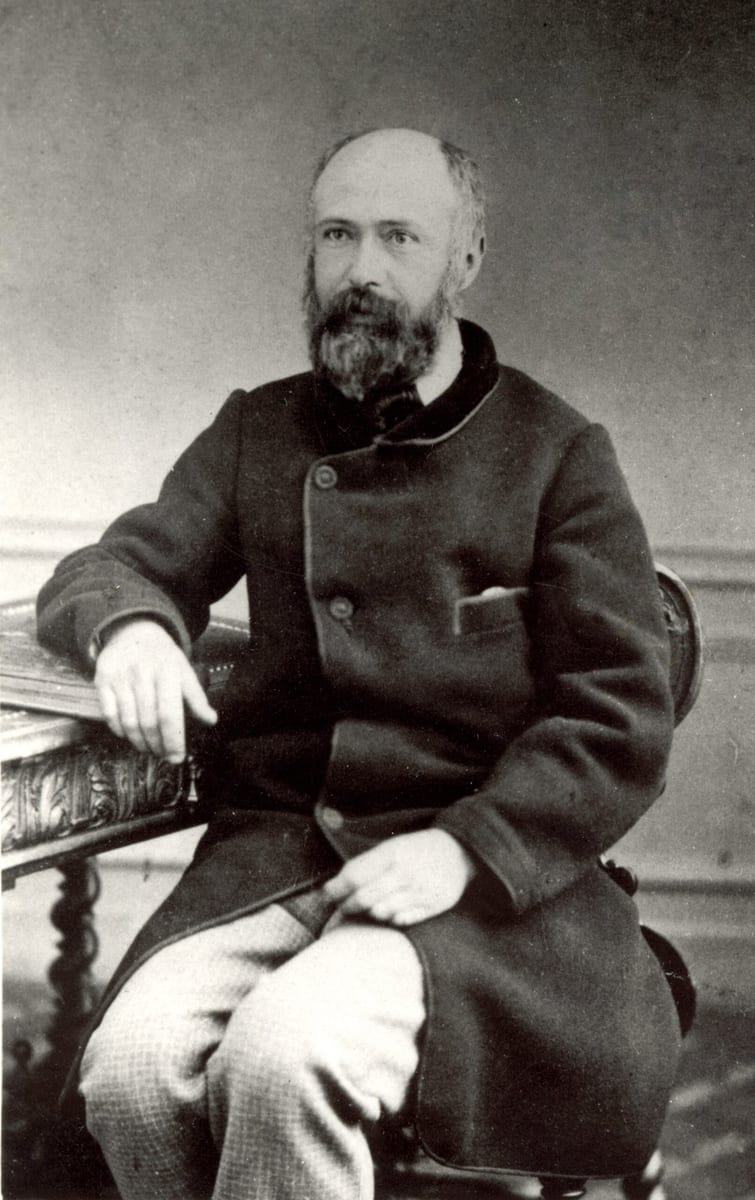 Black-and-white studio photograph of Louis Martin, a balding, bearded man in a dark velvet-collared coat and checked trousers, seated three-quarter-length with one arm resting on a carved table.