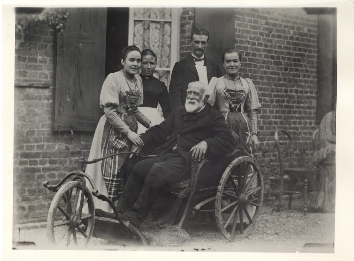 St. Louis Martin seated in a wooden-wheeled invalid chair outdoors against a brick house wall, with his daughters Céline and the Servant of God Léonie Martin and two other people standing beside him.
