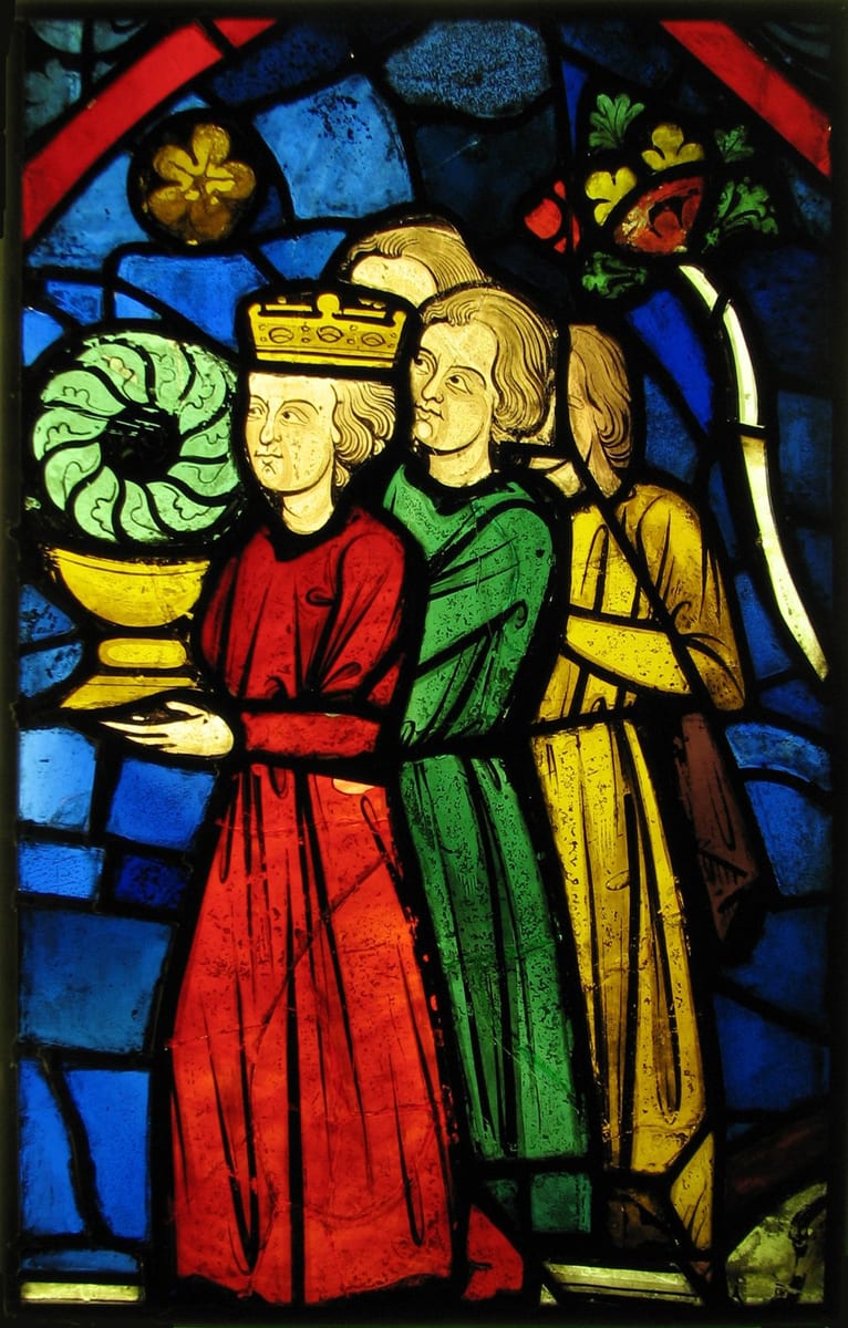 A medieval stained-glass panel of the crowned King Louis IX in a red robe, carrying the Crown of Thorns in a golden cup, with two long-haired attendants in green and yellow behind him against a deep blue ground.