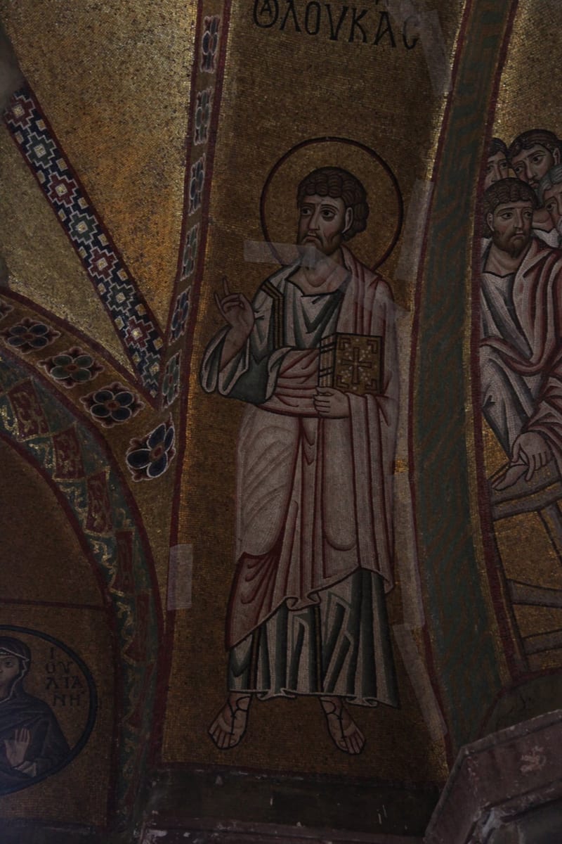 A Byzantine gold mosaic of Saint Luke standing robed with a halo, one hand raised in blessing and the other holding a jeweled, cross-decorated book of his Gospel, with a group of fellow apostles beside him.