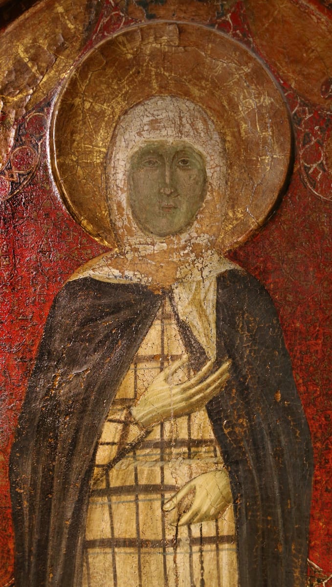 A medieval panel painting of St. Margaret of Cortona standing against a red ground with a gold halo, in a white veil, a dark mantle and a cream tunic crossed with dark squares, her right hand on her breast and a string of prayer-beads in her left.