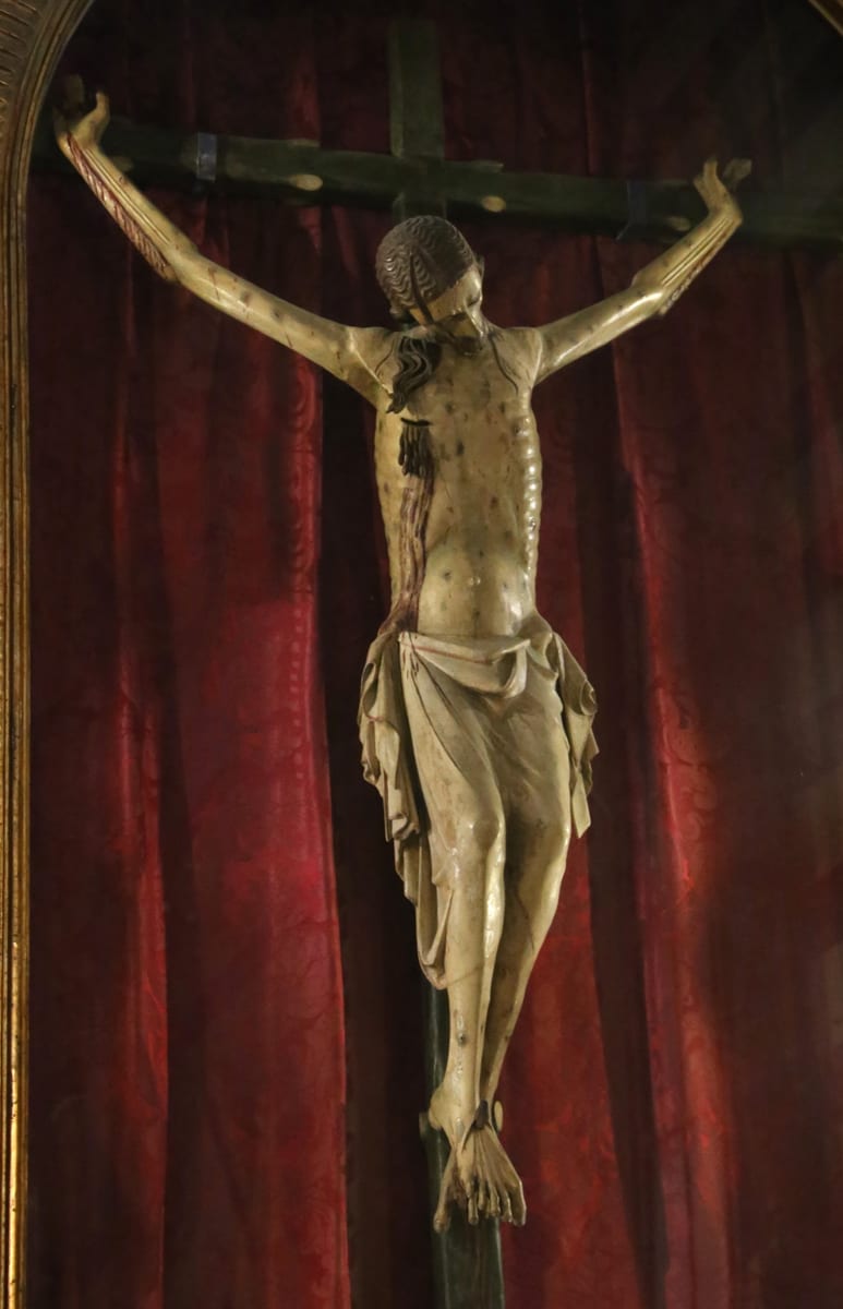 A carved and painted wooden crucifix hangs before a red damask curtain, the dead Christ's thin arms raised in a Y on a dark cross.