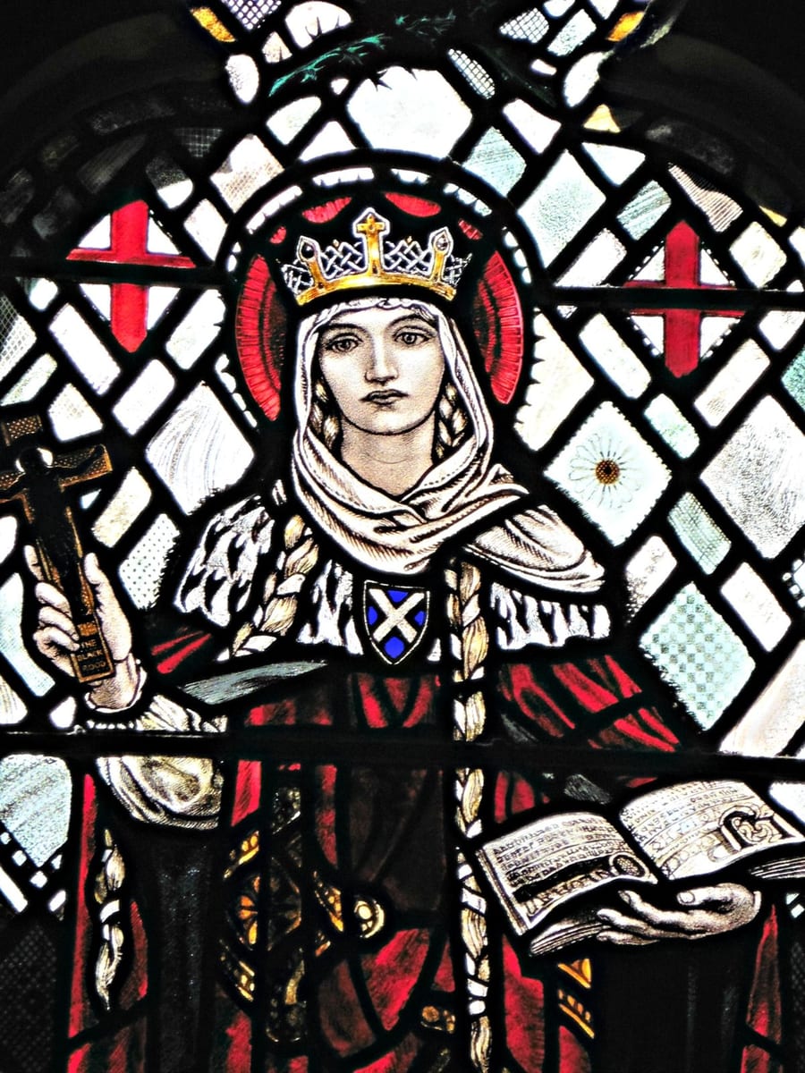 A stained-glass window shows Saint Margaret as a crowned queen with a red halo, a white veil and long blonde braids, holding the Black Rood cross in one hand and an open Gospel book in the other, with a blue shield bearing a white saltire at her breast.