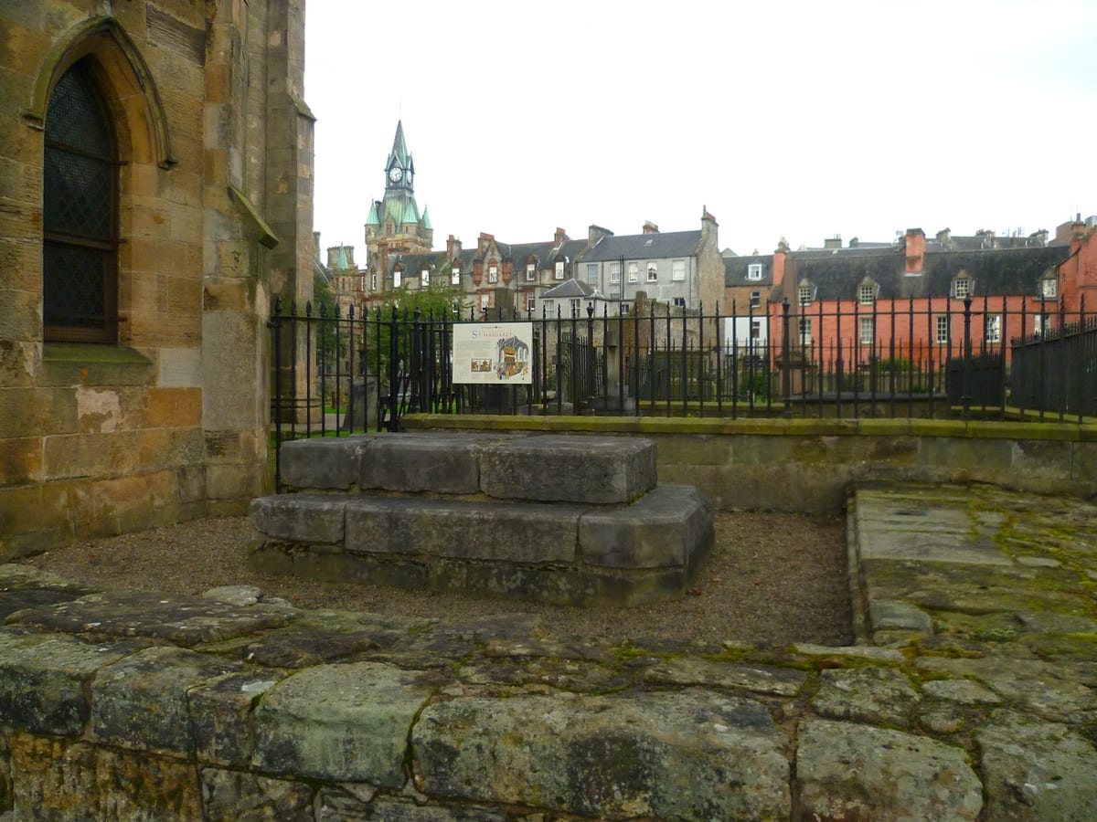 The stepped grey stone base of a shrine stands on gravel beside the buttressed sandstone east wall of Dunfermline Abbey.