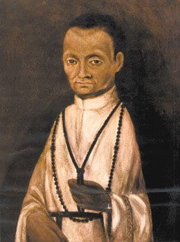 A half-length portrait of a mixed-race Dominican friar in a white habit and black rosary, dark-haired and dark-eyed, looking directly out at the viewer against a plain dark background.