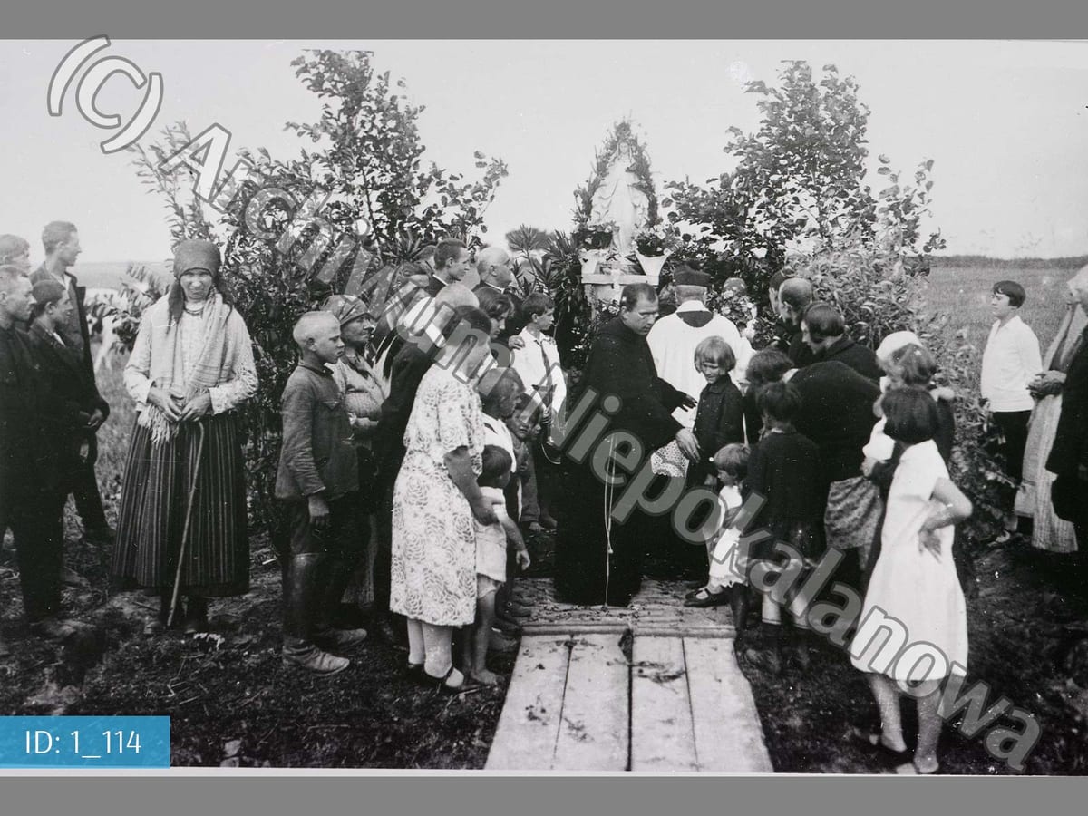 A friar arranges village children into their places at the blessing of a statue of the Immaculata standing on an open field.