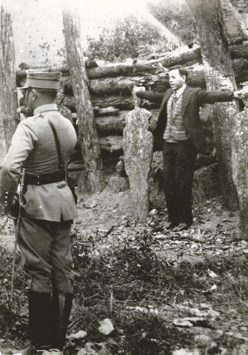 Father Pro stands before a log stockade with his arms stretched straight out, while an officer with a sword watches him from the foreground with his back to the camera.