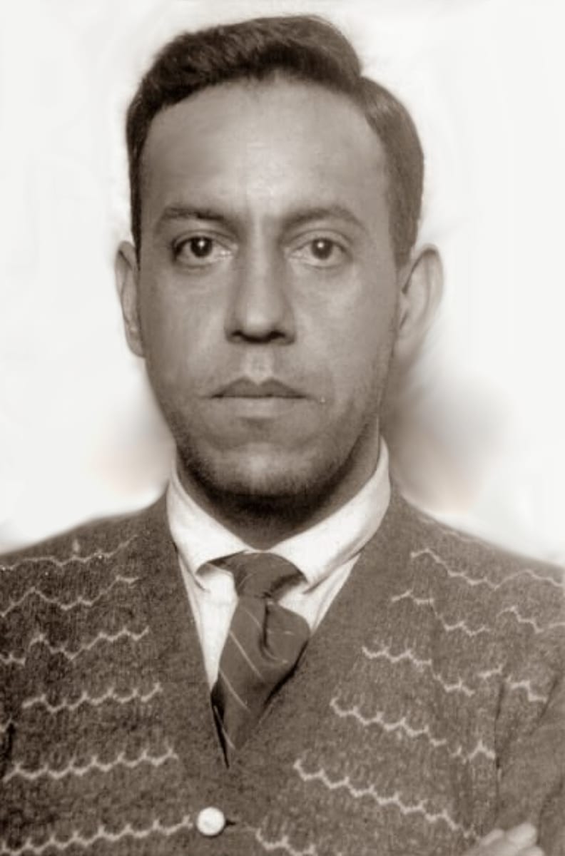 Father Pro looks straight into the camera with his arms folded, in a light shirt, striped tie and a patterned cardigan.