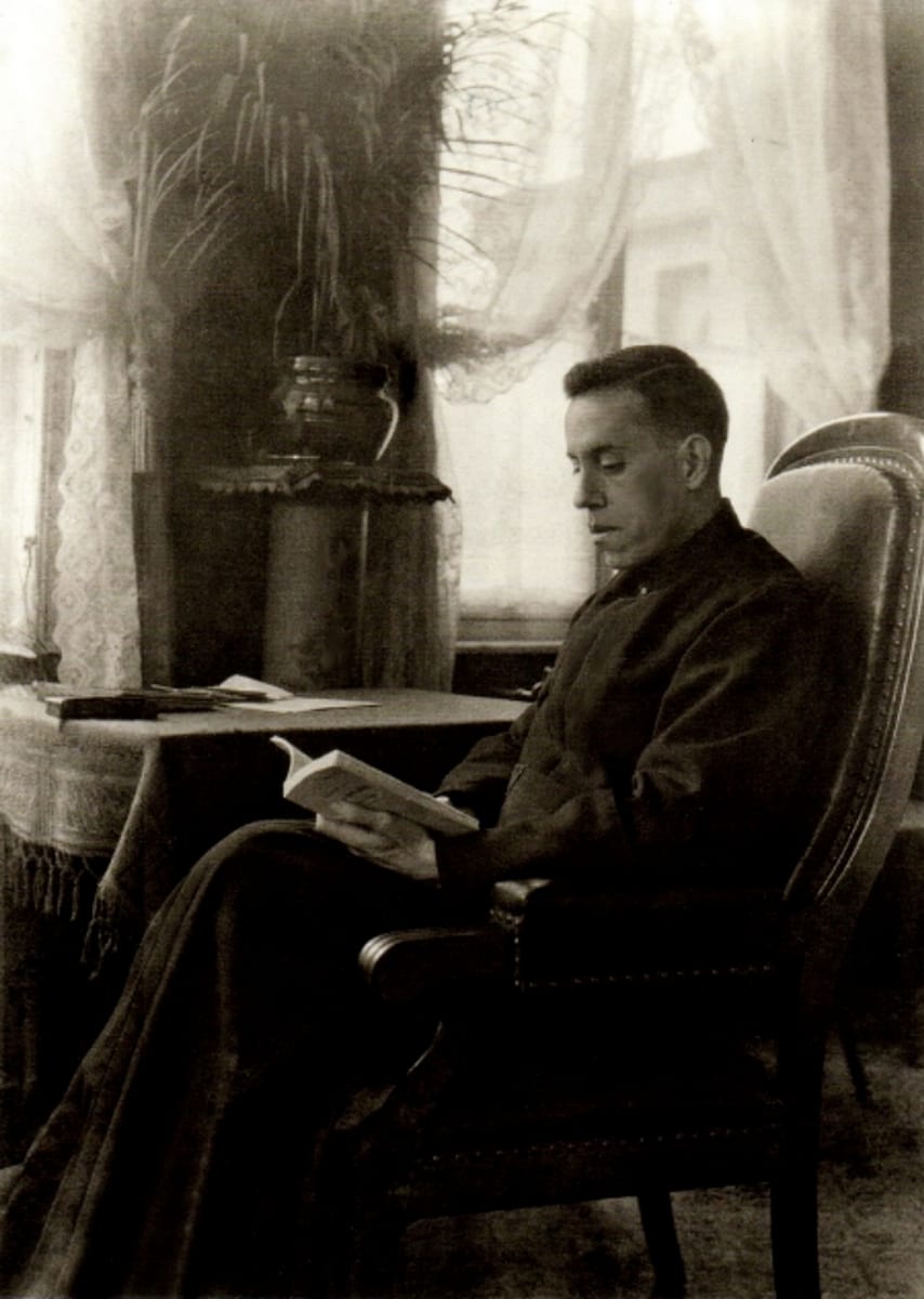 Father Pro sits in profile in a high-backed armchair by a curtained window, a book open on his lap, wearing a black cassock.