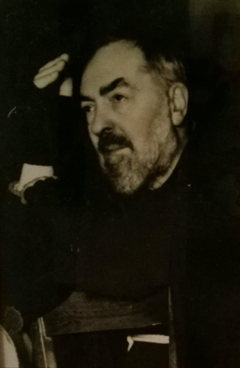 A black-and-white photograph of an elderly bearded Capuchin friar in a dark habit, seated and turned in profile with one hand raised, holding papers, in a dimly lit room.