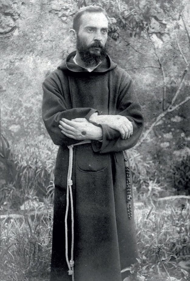 A black-and-white photograph of a young, bearded St. Pio standing in his Capuchin habit with the wounds in his hands shown.