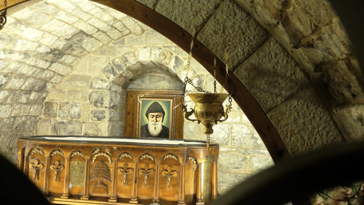 A low carved wooden tomb under a stone vault, with a brass lamp hanging above it and a framed portrait of the saint on the wall behind.