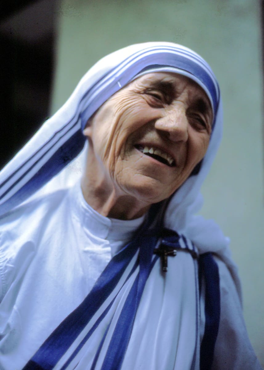 An elderly Mother Teresa, in her white sari with blue border and a small crucifix pinned at the shoulder, laughs with her head tilted back.