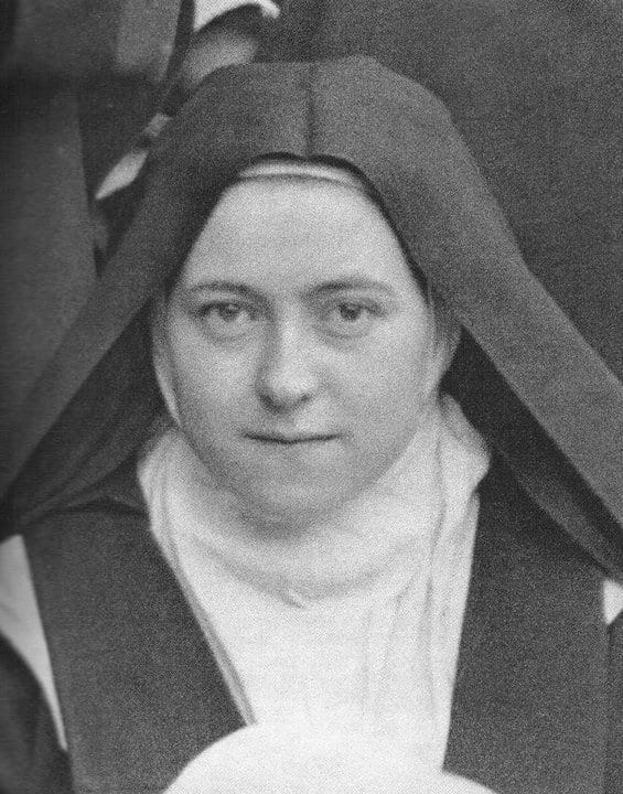 A close-up photographic portrait of a young Carmelite nun, Saint Thérèse of Lisieux, wearing the order's dark habit and veil over a white wimple, looking toward the camera with a calm, faint smile, photographed in the courtyard of the Carmel of Lisieux.
