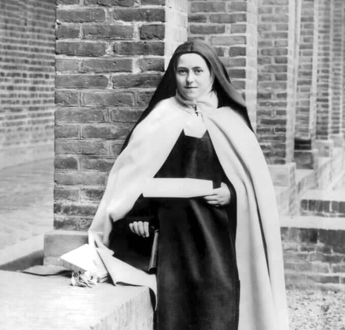 St. Thérèse standing full-length against the courtyard wall of the Carmel of Lisieux, holding a parchment in one hand and a book on the Su-tchuen mission in the other, photographed after July 3, 1896 AD.